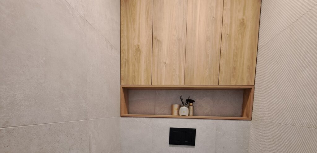 Elegantní vestavěná skřínka do niky na toaletě byla vyrobena na míru do novostavby. Skřínka se skládá z uzavřené a otevřené části. Uzavřená část je včetně dvířek s bezúchytkovým systémem otevírání. Vše je vyrobeno v odstínu dřeva, které kontrastuje s obkladem místnosti.