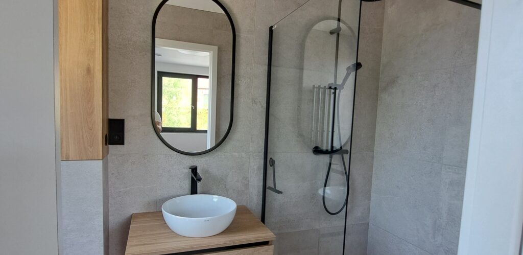 Minimalistické skřínky do koupelny jsou navrženy v odstínu dřeva, který koresponduje s obkladem koupelny. Skřínky jsou vybaveny moderním bezúchytkovým systémem.